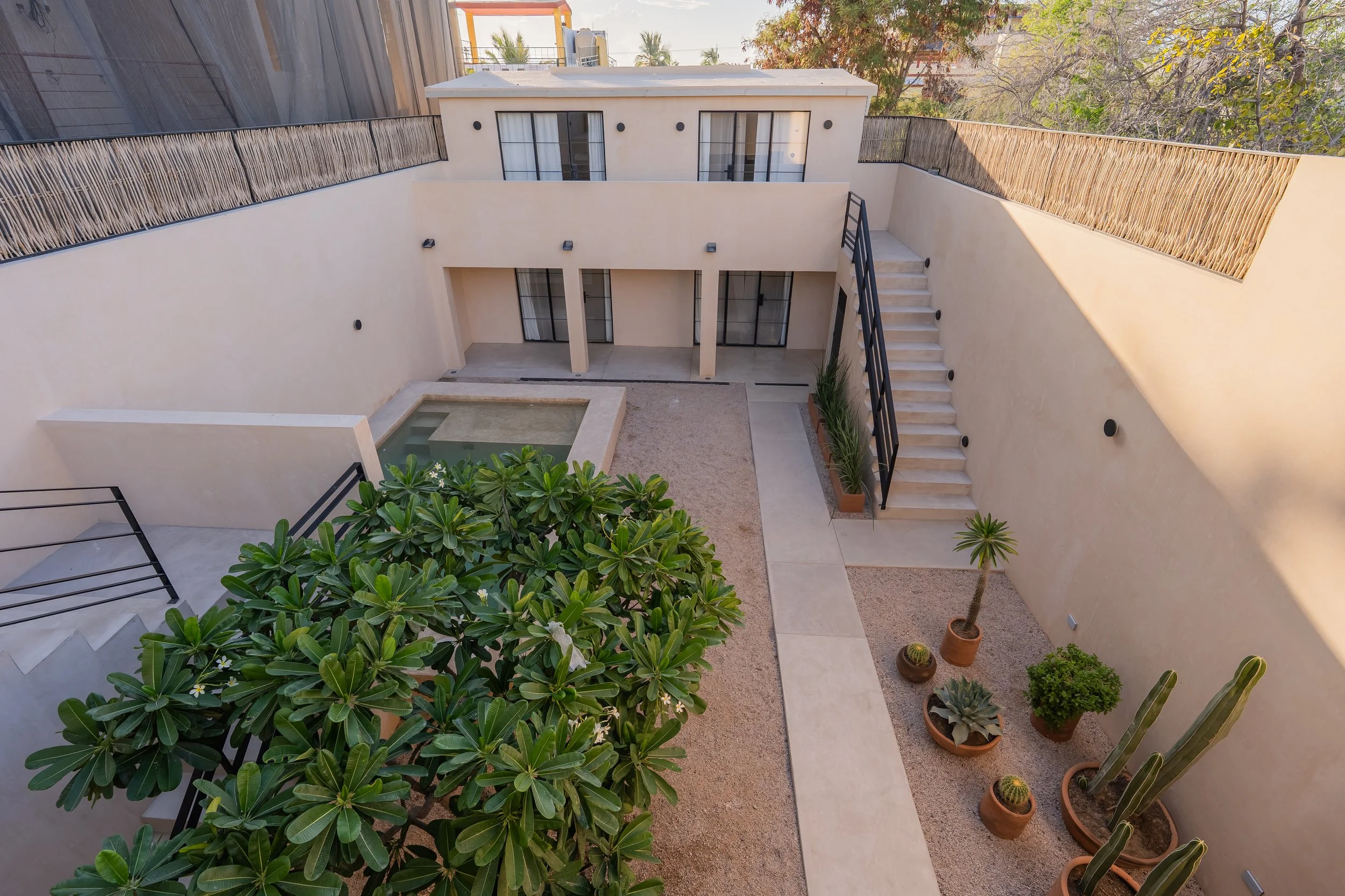 A modern, two-story residential building with sliding glass doors and windows, surrounded by a courtyard with potted plants, a small pool, and a staircase on the right side.