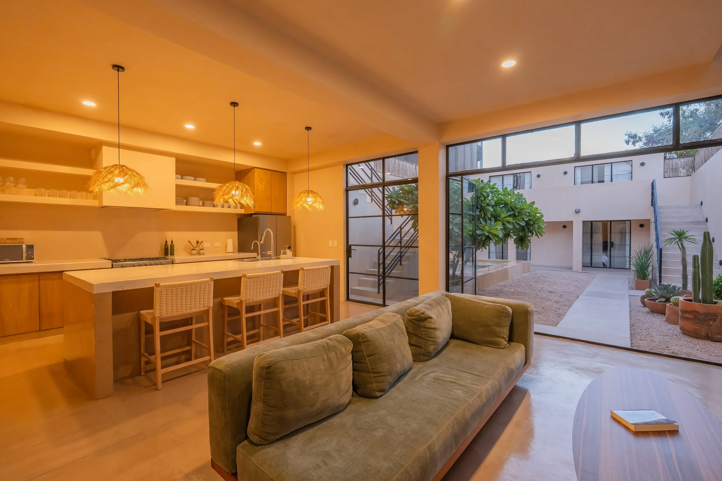 Open-plan living area with a kitchen island, seating, and large glass doors opening to an outdoor patio with plants and staircase, illuminated by warm lighting.