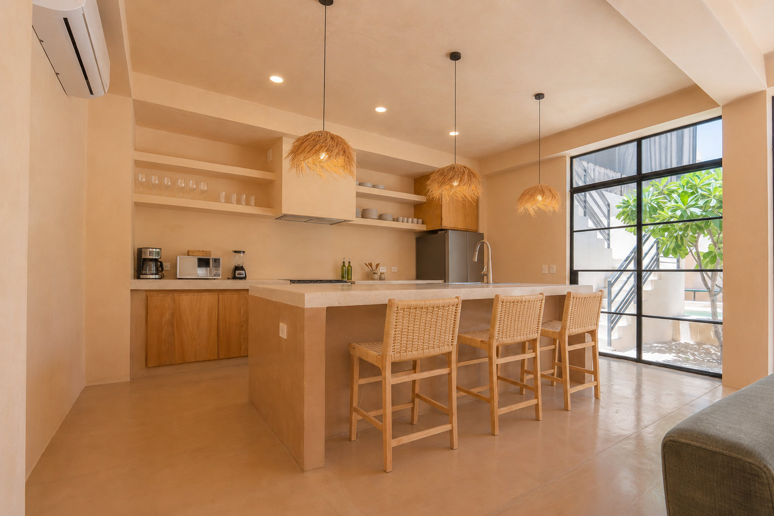 Modern kitchen with beige walls, wooden cabinetry, a large island with seating, pendant lights with natural fibers, and a large window showing a tree outside.
