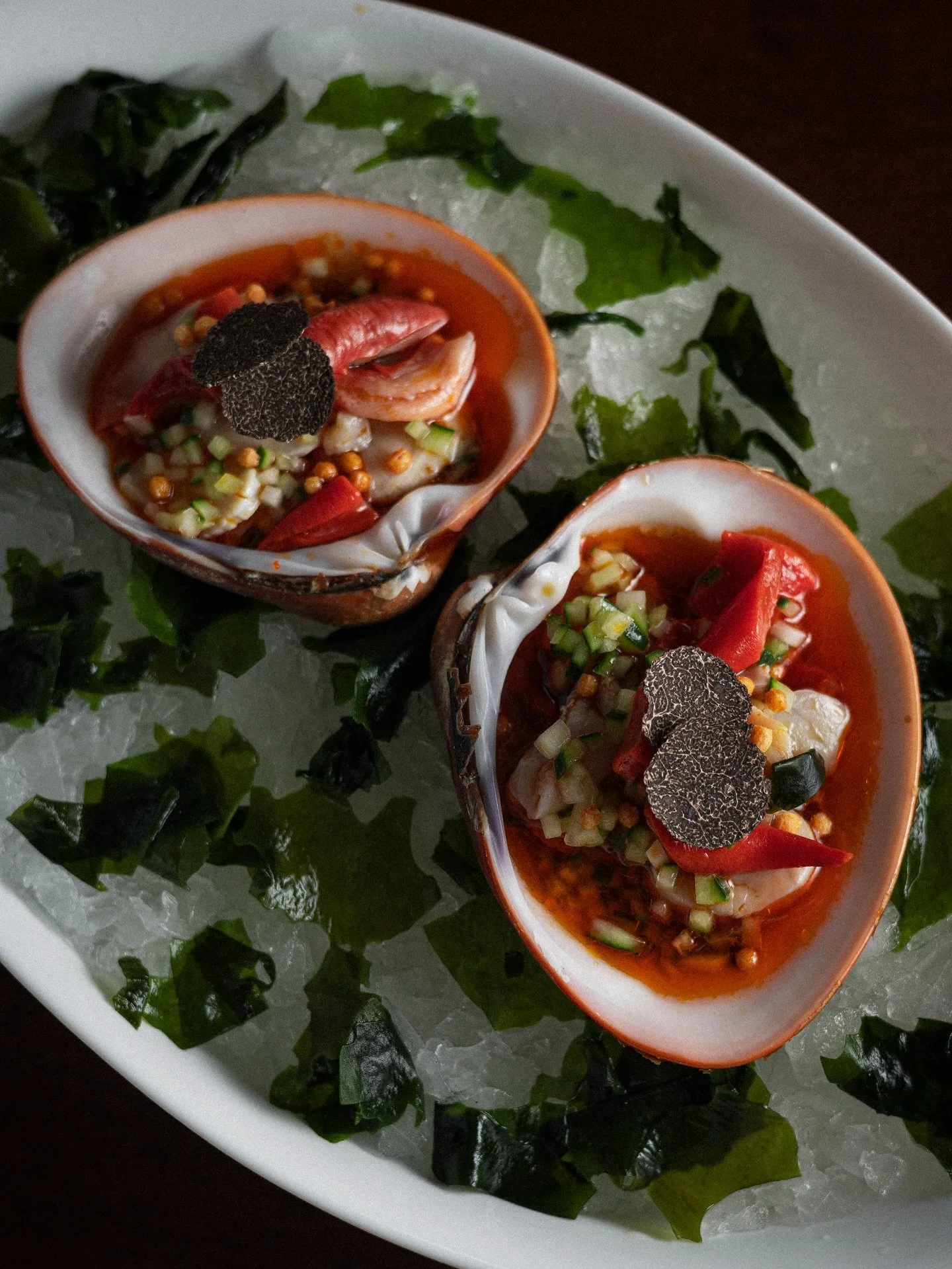 Shells filled with seafood and topped with black truffle slices, served on a bed of ice with green seaweed garnishes.