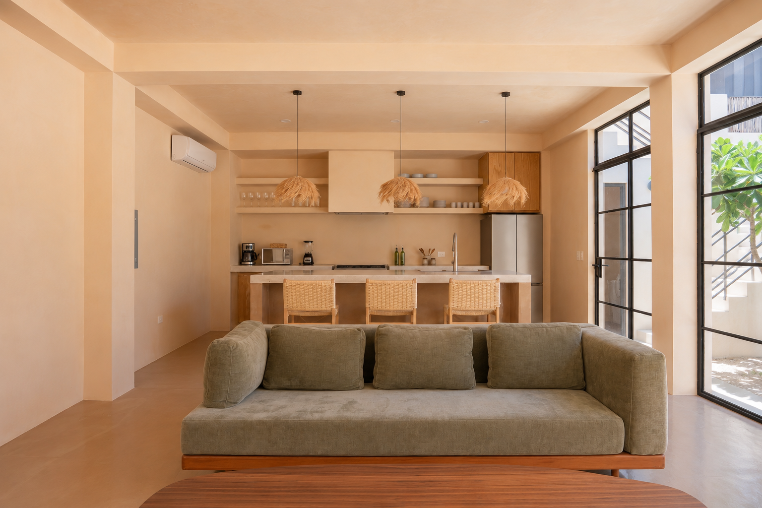 Living room with green sofa, open kitchen with beige cabinets, pendant lights, bar stools, large windows with view of outdoor stairs and greenery.