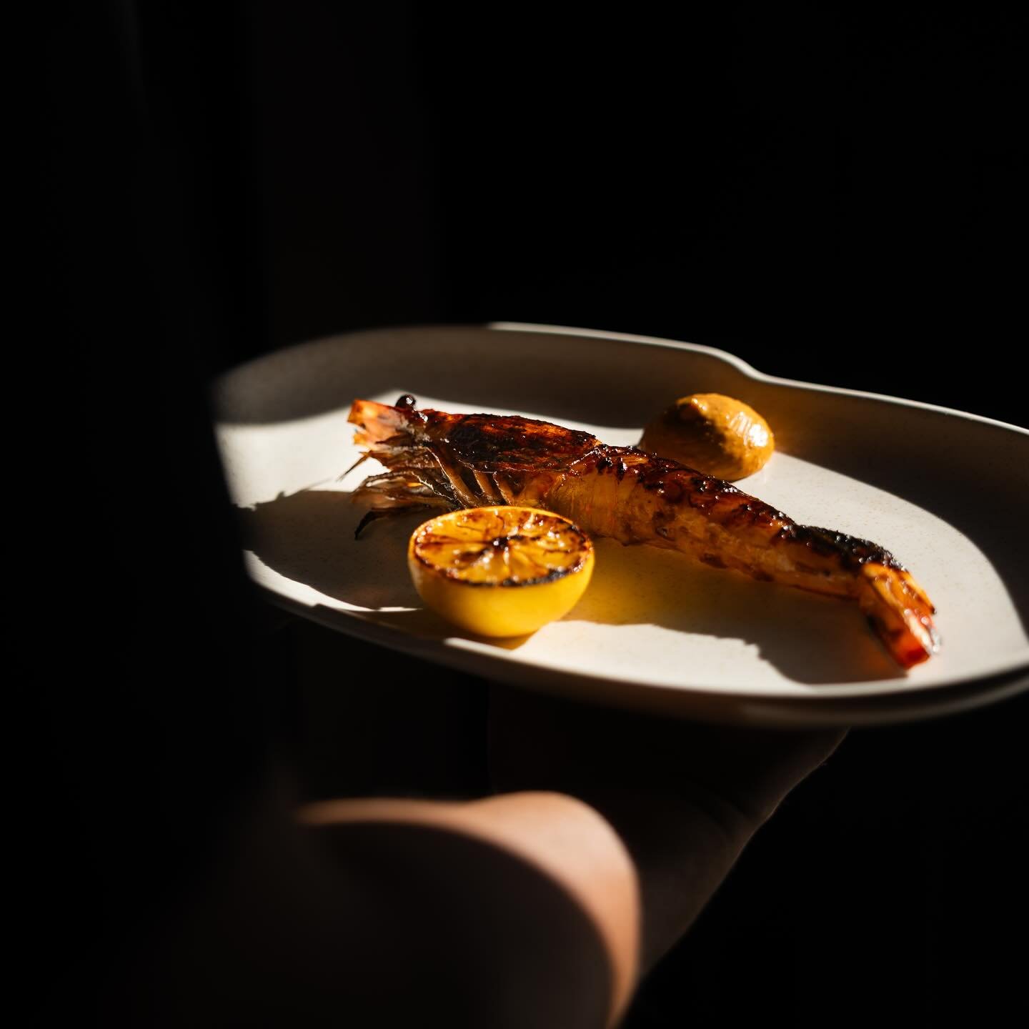 Grilled shrimp on a white plate with grilled lemon halves and a small round scoop of sauce, illuminated by a focused light in a dark setting.