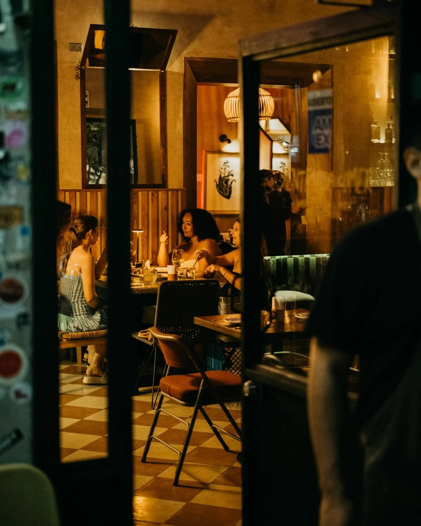 People sitting at a cozy restaurant table, seen through a window, with warm lighting and wooden decor.