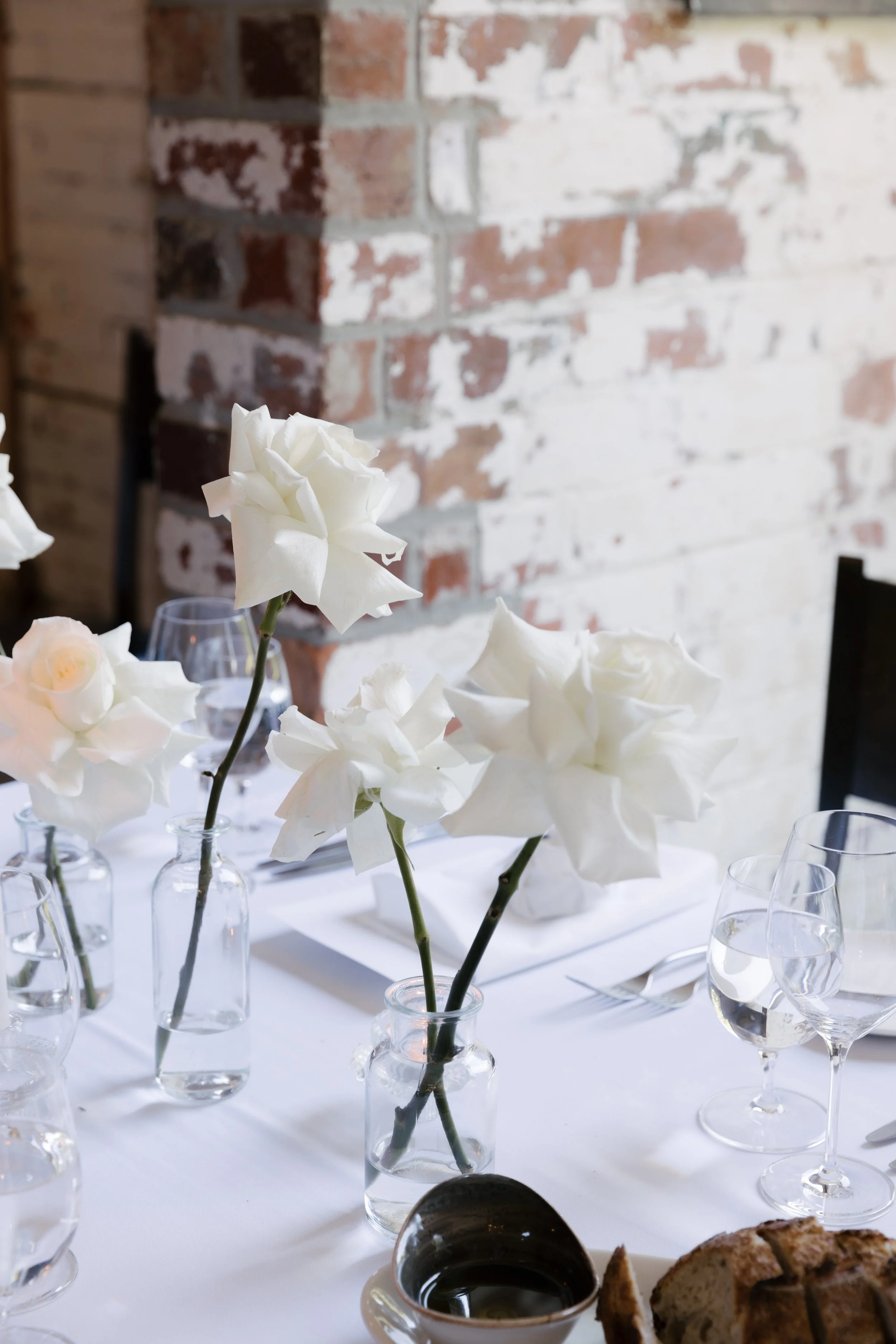 A dining table decorated with white flowers in small glass vases, wine glasses, a bread roll, and a small dish of sauce, with a exposed brick wall in the background.