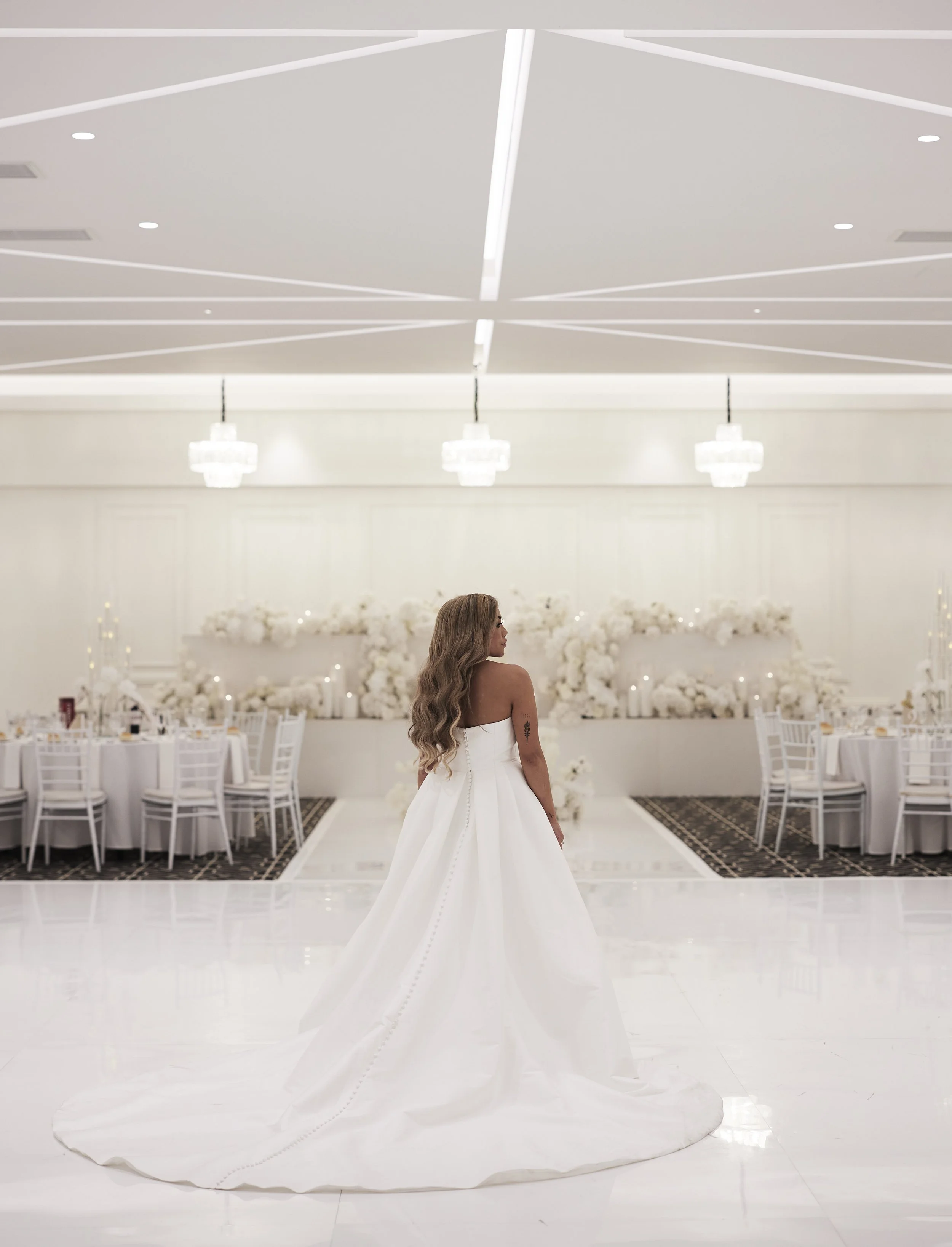 A bride in a white wedding dress with long hair stands in an elegant white hall decorated for a wedding reception.