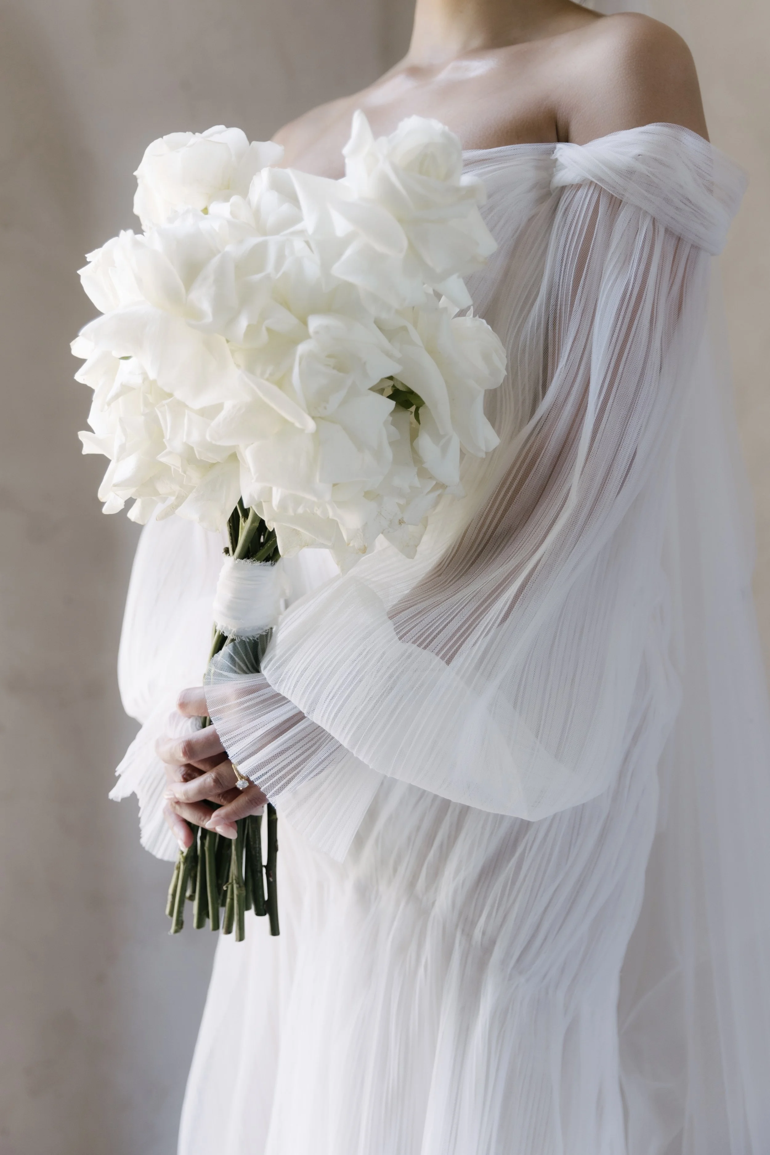 A woman dressed in a flowing white gown holding a bouquet of white flowers, standing against a neutral background.