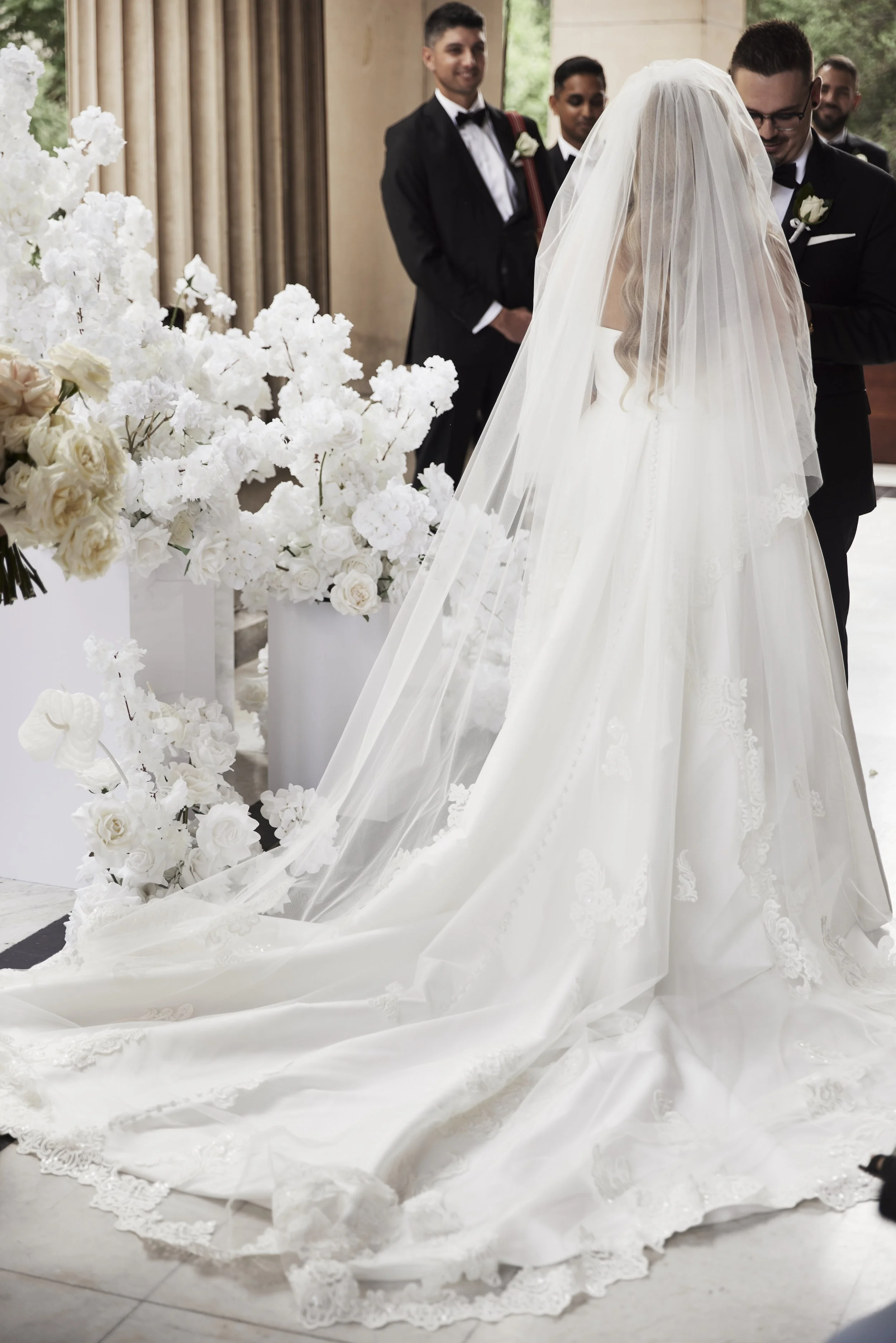 A bride in a white wedding gown with a long train and veil is at her wedding ceremony. She is surrounded by white flowers, and a groom and groomsmen dressed in tuxedos are present, smiling and looking at her.