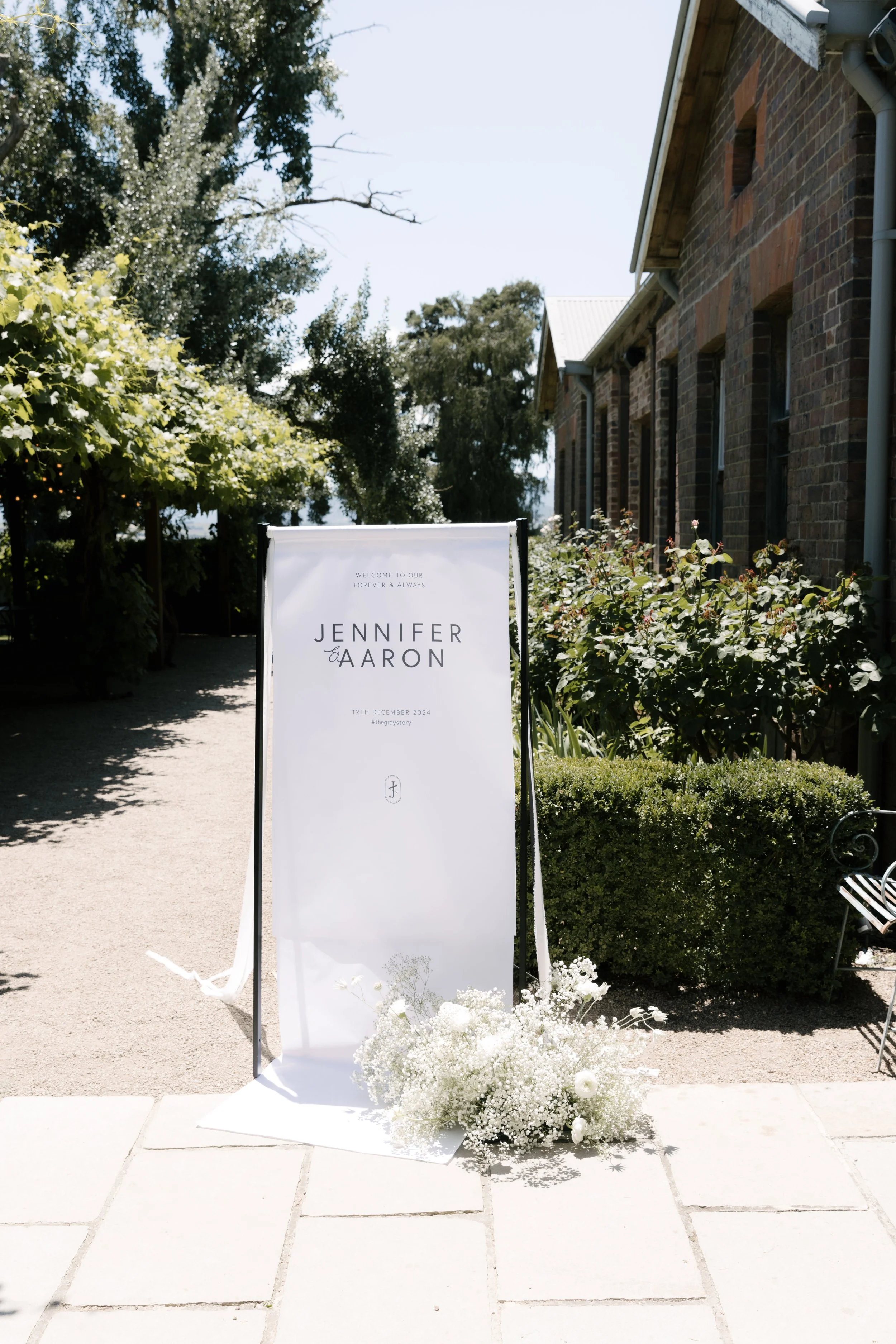 White wedding sign with black text reading 'Jennifer & Aaron' and the date '12th December 2024,' decorated with white flowers at the base, placed in front of a brick building surrounded by greenery on a sunny day.