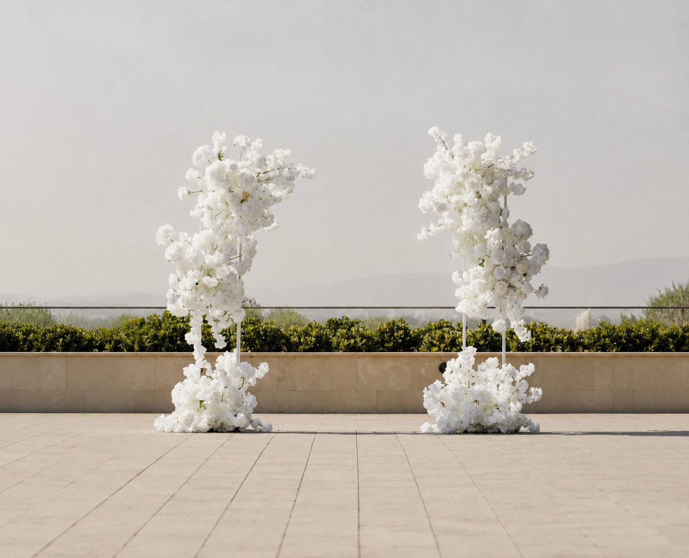 Two tall abstract sculptures made of white flowers on thin white stands, placed on a paved outdoor area with green shrubbery and a hazy sky in the background.