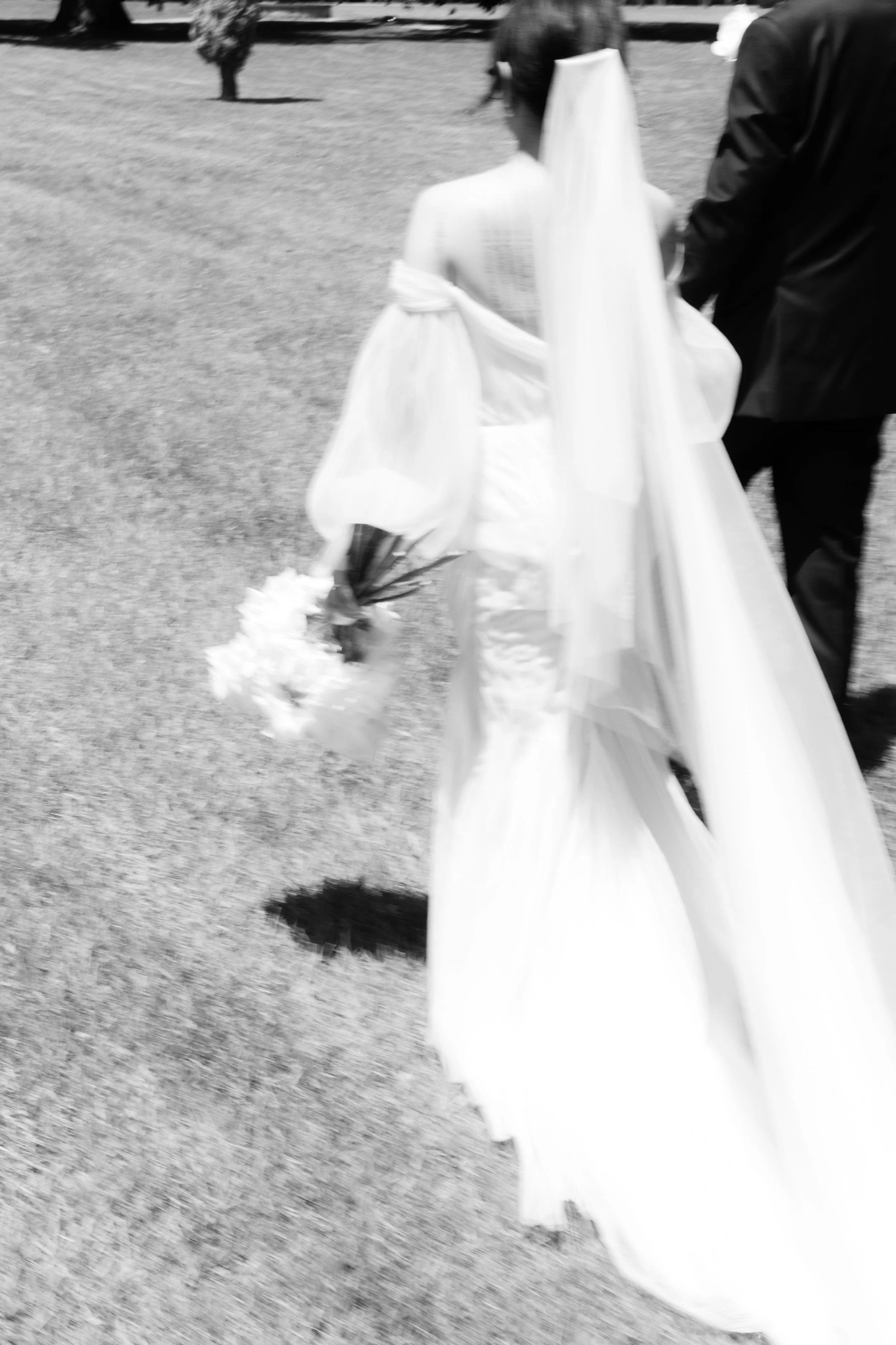 Black and white photo of a bride and groom walking outside. The bride wears a white dress and headpiece, carrying a bouquet, and the groom is in a dark suit.