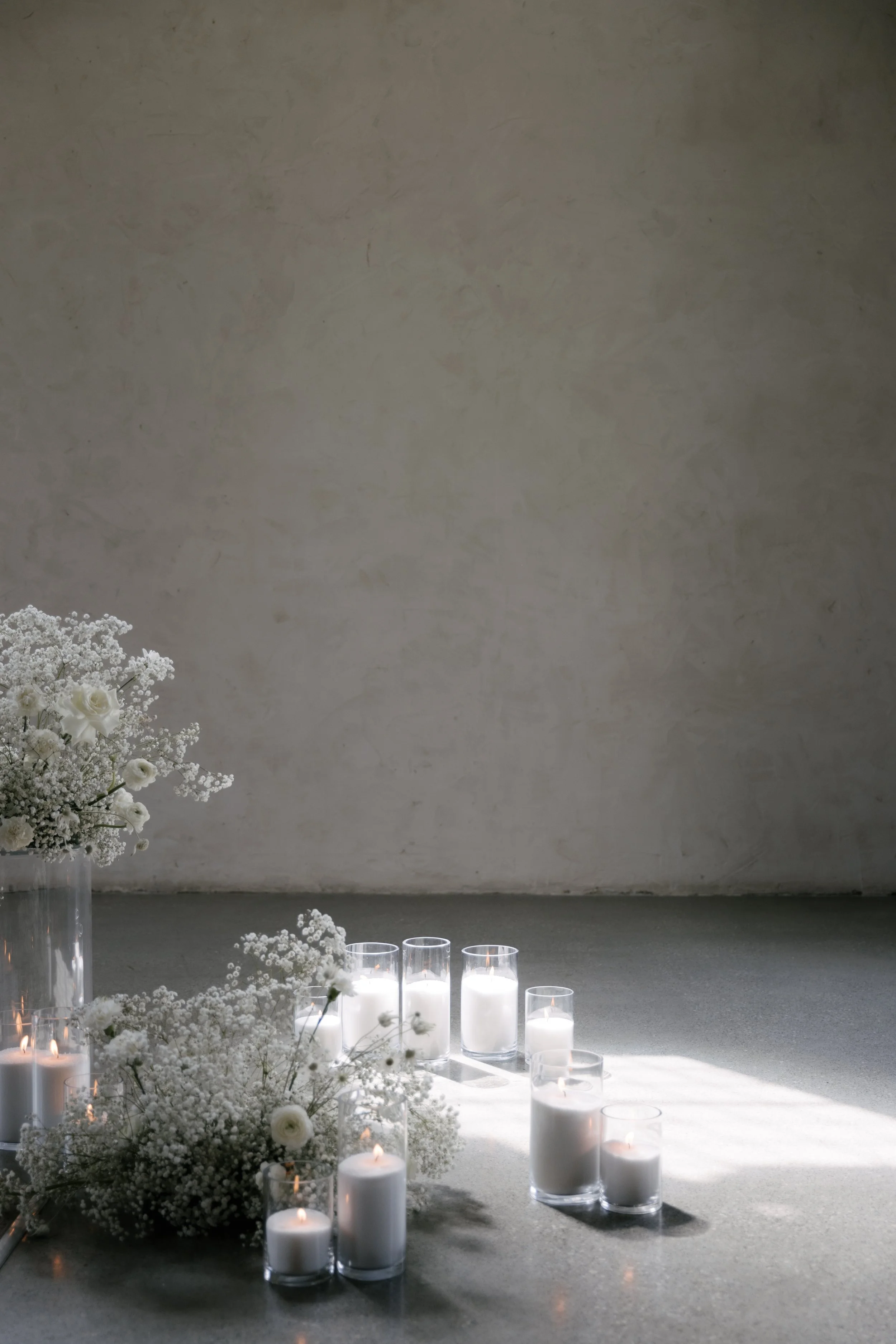 White floral centerpiece with baby's breath and roses in a clear vase, surrounded by lit white candles in glass holders on a flat surface, against a textured beige wall.
