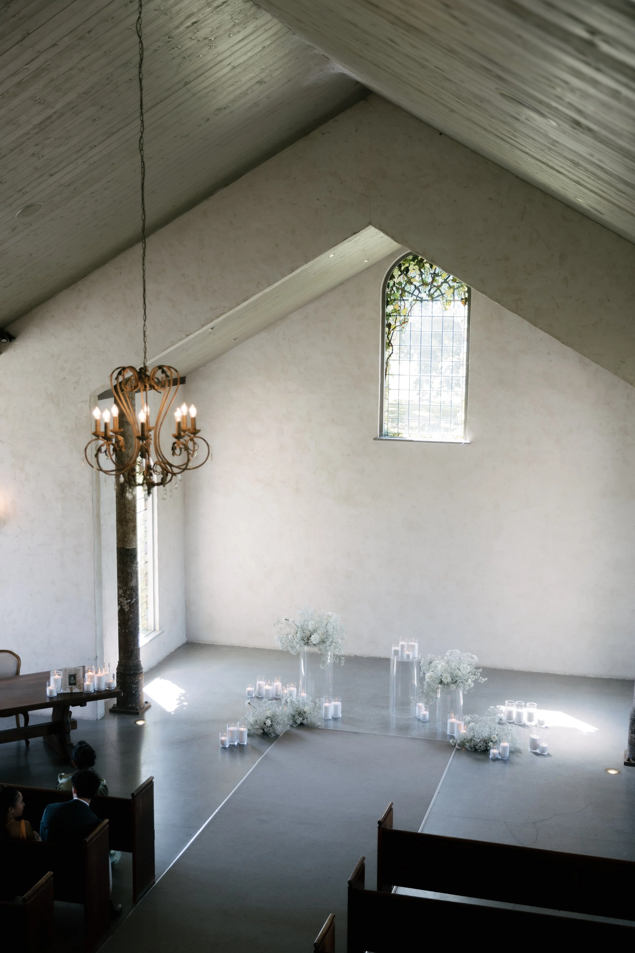 Interior of a church or wedding venue with white walls, high vaulted ceiling with wooden panels, a chandelier hanging from the ceiling, a tall stained glass window with greenery outside, and floral arrangements with candles on the floor.