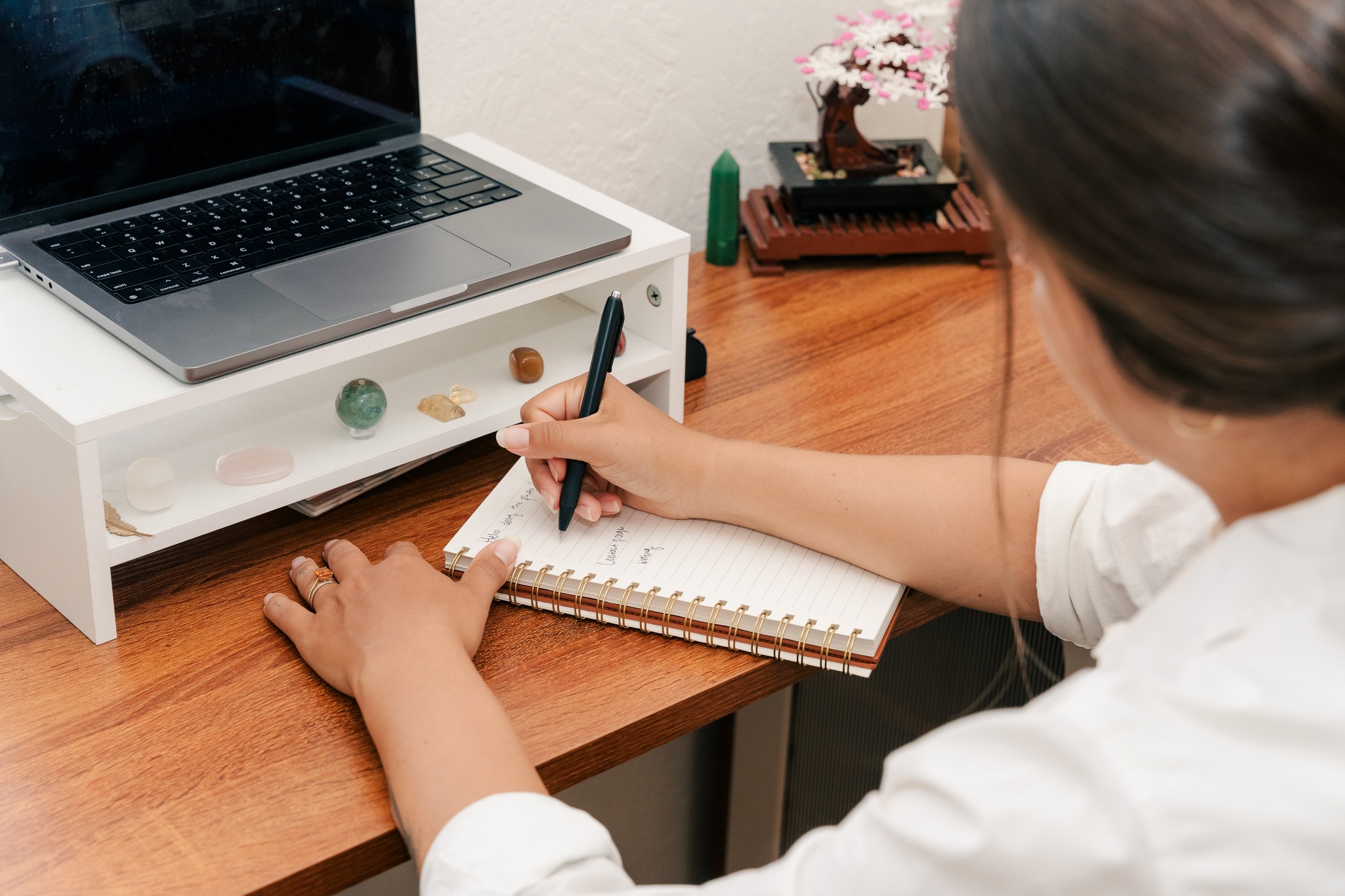A person sitting at a wooden desk writing in a spiral notebook with a black pen. On the desk, there is a white shelf holding various polished stones and a laptop. In the background, a decorative pink and white flower tree is visible.