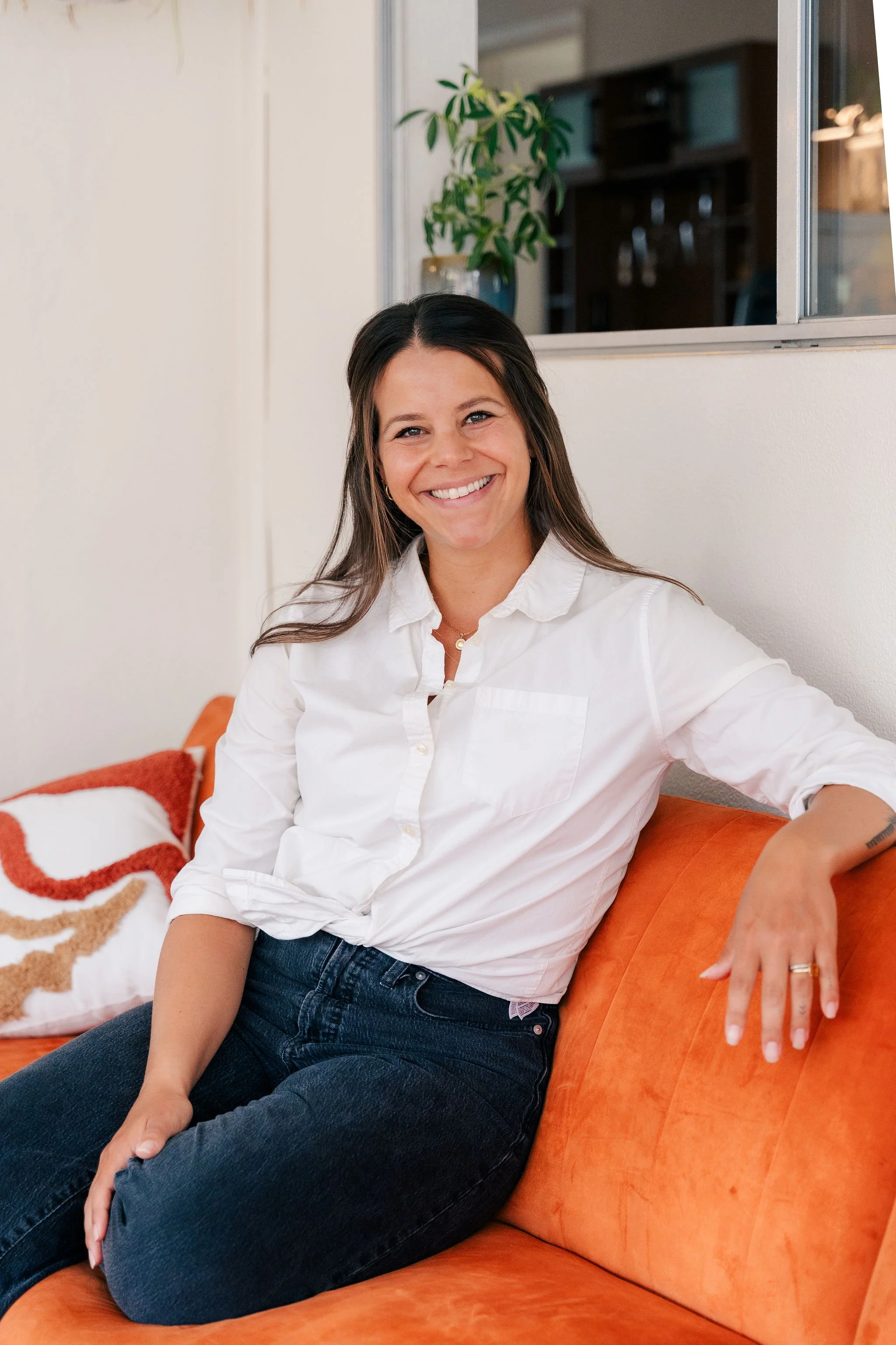 A woman with long brown hair smiling, seated on an orange couch in a room with white walls, wearing a white shirt and dark jeans.