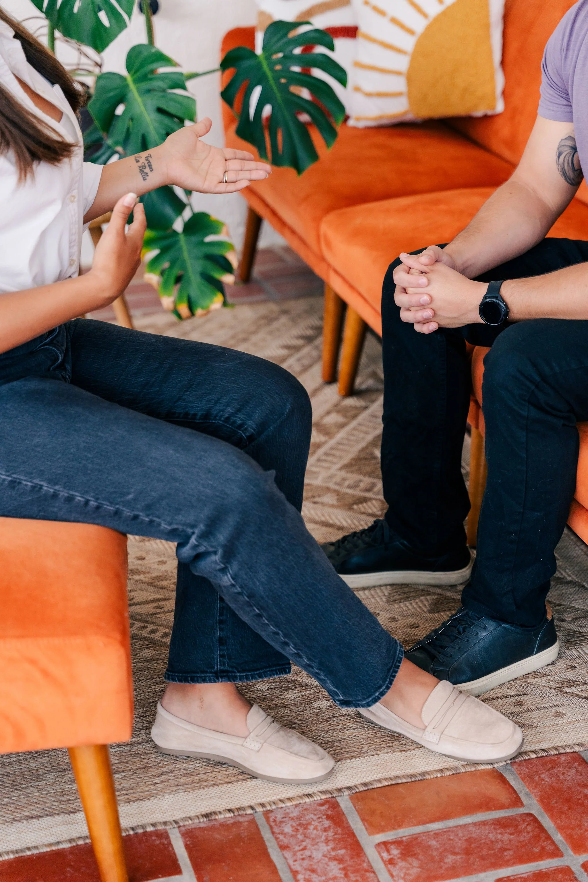 Two people sitting on an orange sofa in a living room, engaging in conversation. One person's hands are visible, move in a gesture, with a tattoo. The other person, wearing a light purple shirt and a wristwatch, has hands clasped together. There are large green monstera leaves and decorative pillows with orange and white patterns in the background, on a terracotta tiled floor.