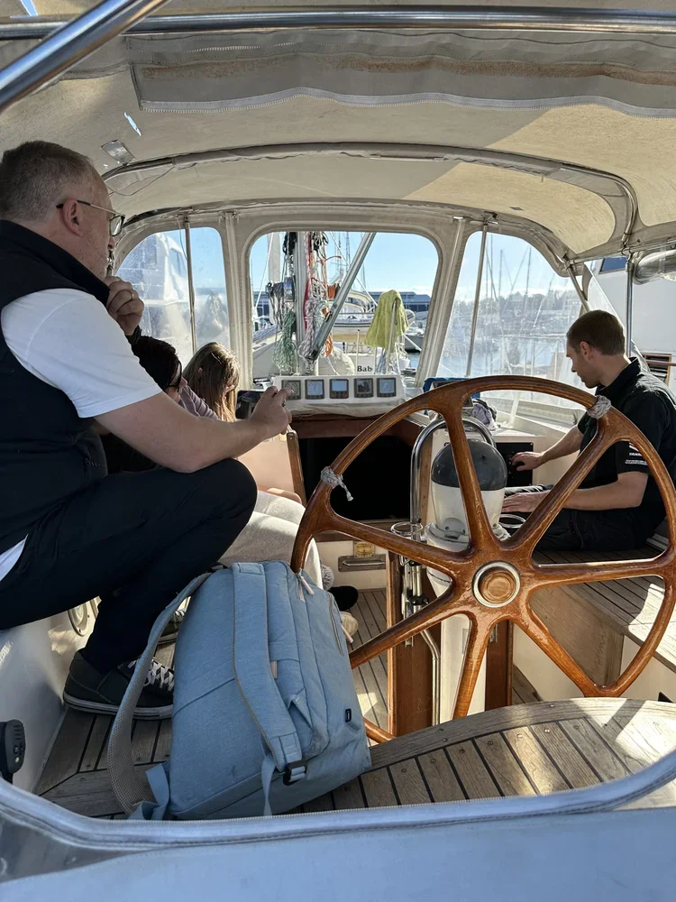 People sitting inside a boat on a sunny day, with a large wooden steering wheel and various navigational instruments at the front, parked at a marina with other boats in the background.