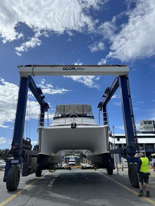 A large boat is being lifted by a boat lift at a marina on a sunny day with a bright blue sky and scattered clouds.