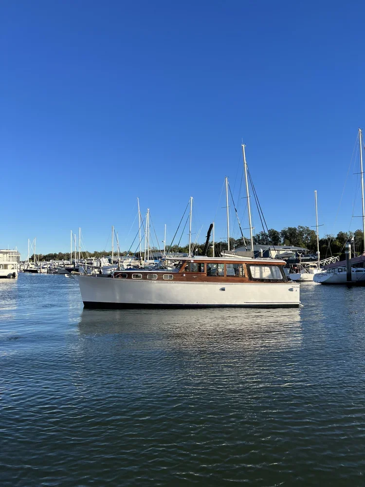 A white and brown motor yacht sailing on calm water in a marina with multiple sailboats docked on both sides and a clear blue sky above.