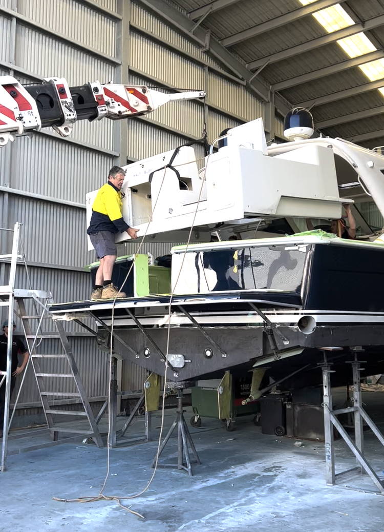 A man in a yellow and black shirt standing on the deck of a boat, working on the white superstructure of a helicopter mounted on the boat within a boat garage or maintenance facility.