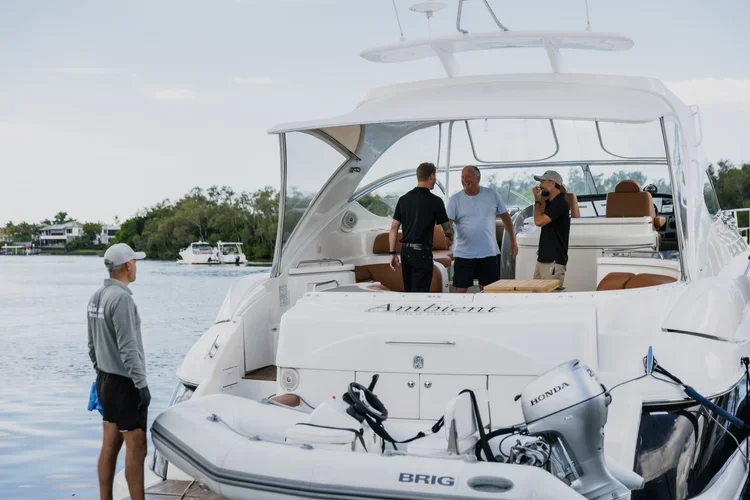 Four men on a luxurious white yacht named 'Ambient' docked on the water, with two others on the dock nearby, and a calm body of water with other boats in the background.