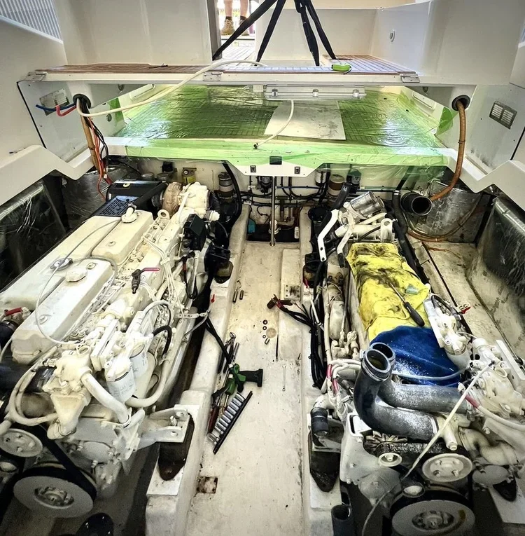 Interior of a boat or yacht engine room with two large diesel engines, various parts, tools, and equipment, and a glass-covered compartment at the top with green support.