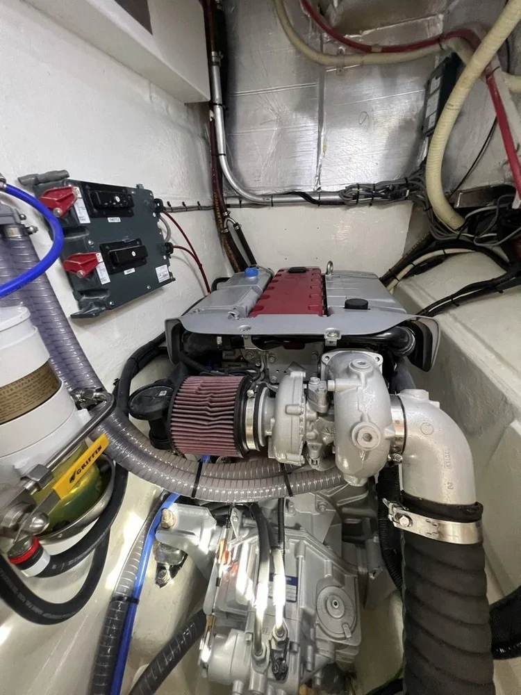 A small engine with a red valve cover, air filter, vacuum hoses, and electrical components in a confined space, likely inside a boat or vehicle engine compartment.