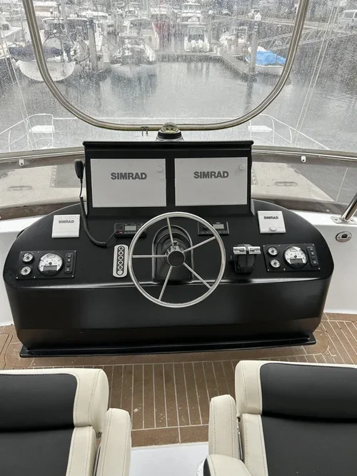The image shows the captain's control panel on a boat, featuring a steering wheel, two Simrad navigation screens, and various gauges and controls, with boat docks visible outside the windows.
