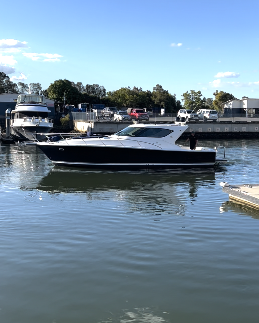 A black and white motor yacht docked in a marina, with other boats and cars in the background under a clear blue sky.