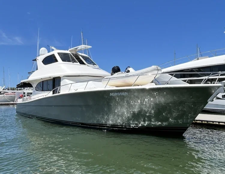 A large white motor yacht anchored at a marina with other boats in the background, under a clear blue sky.
