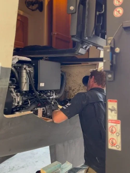 A mechanic working inside a vehicle's engine compartment, wearing a black shirt and cap.
