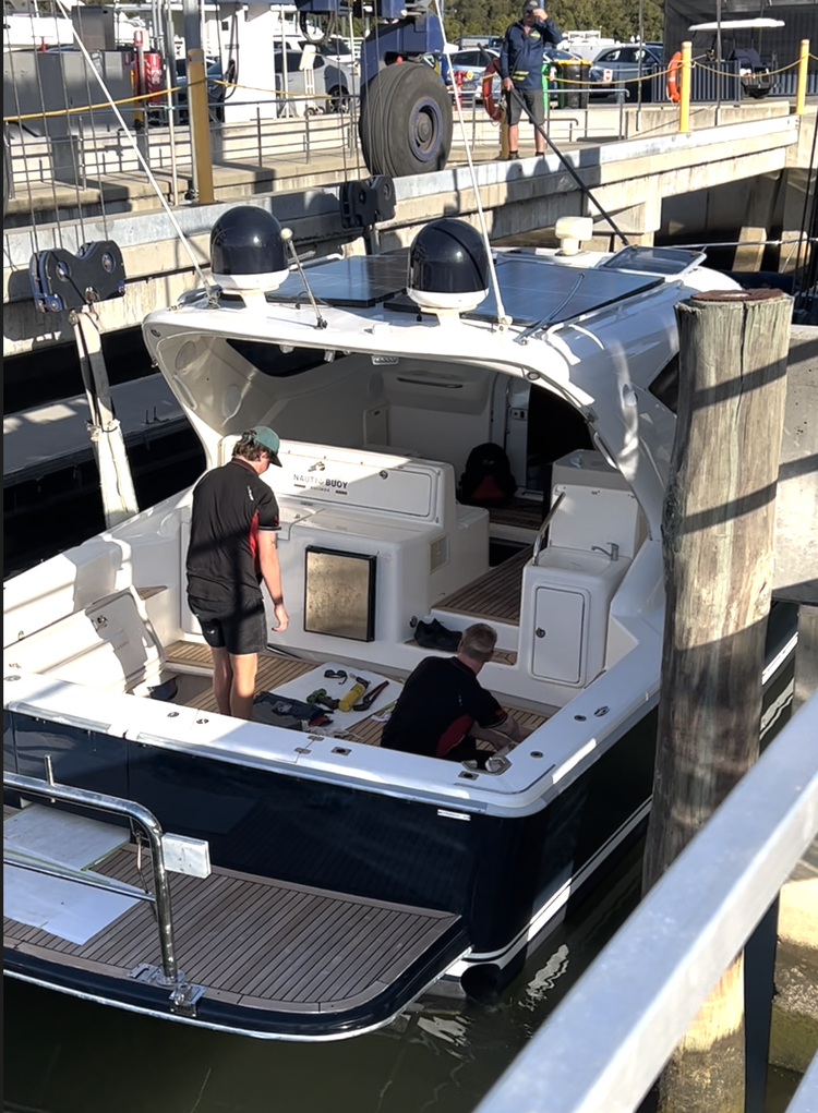 Two men working on a white motorboat docked at a marina, with tools on the deck and other boats and vehicles visible in the background.