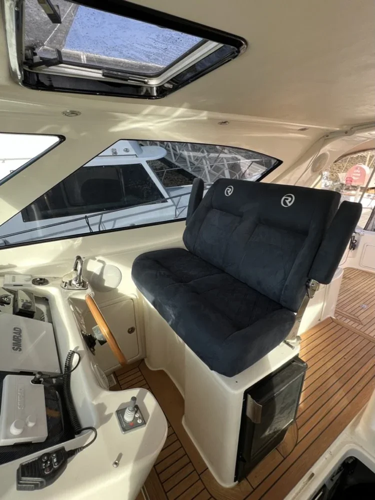 The image shows the helm of a boat with a black cushioned captain's seat, a wooden steering wheel, and various navigational equipment, with a large window and a hatch overhead.