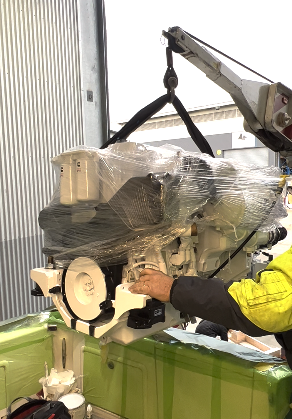 A person working on a large, white machinery part wrapped in plastic, with a crane lifting it, in an industrial setting.