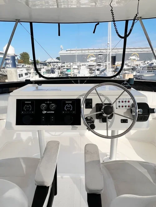 View of a boat's cockpit with navigational controls, steering wheel, and electronic displays, overlooking a marina with docked boats and a yacht in the background.
