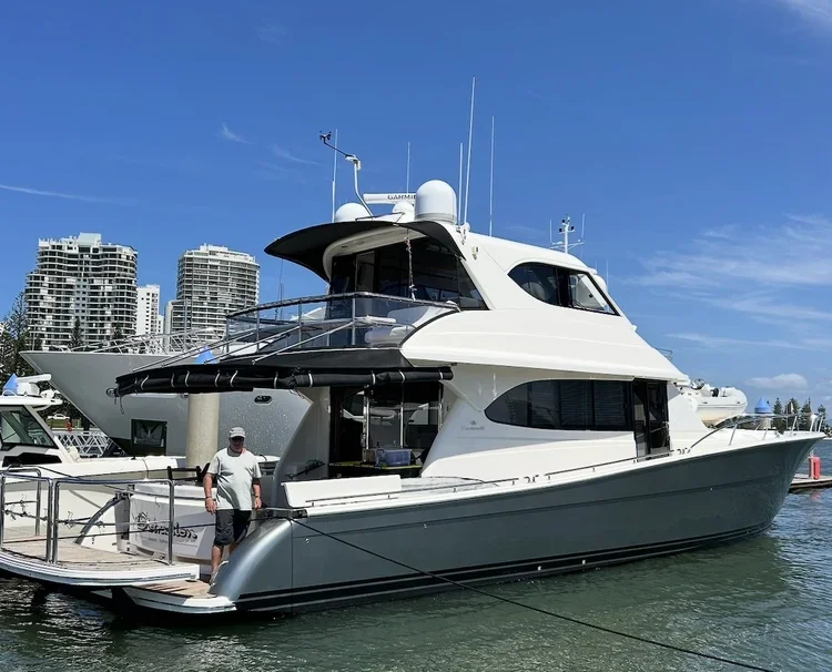 A white yacht docked in a marina with a city skyline in the background, a man standing on the deck.