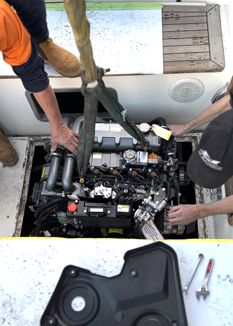 Two workers install or service a boat engine through an open access point in the boat's deck. One worker is wearing an orange and black shirt and brown boots, the other a black cap and black shirt. Tools and a black cover are visible nearby.