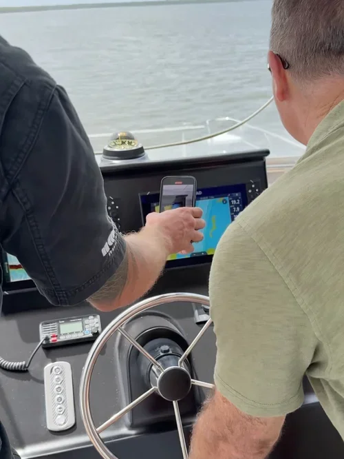 Two men on a boat, one showing the other a smartphone with a sonar or fish finder display, near a steering wheel and electronic equipment, on a body of water.
