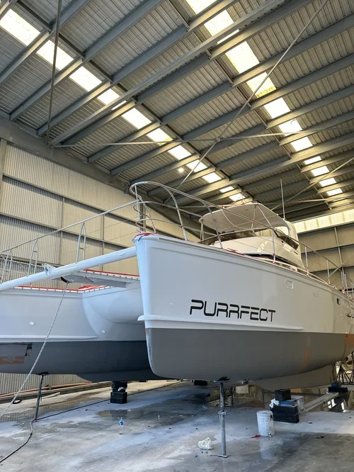 A white sailboat named 'Purrfect' inside a large industrial warehouse, elevated on supports for maintenance or storage.