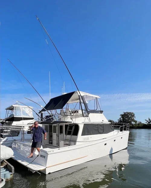 A man standing on a white motor yacht docked at a marina during daytime with clear blue skies.