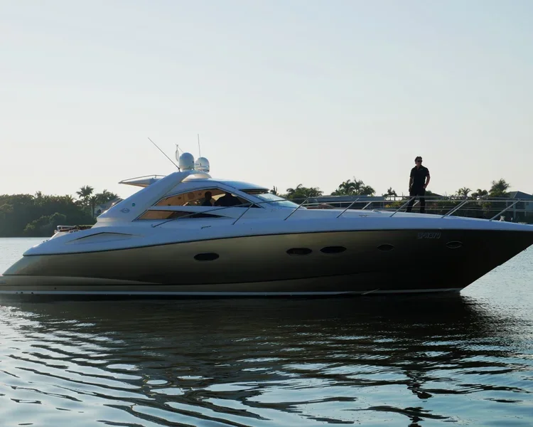 A luxury yacht on calm water with a person standing on the bow, and another person inside the boat, with trees in the background.