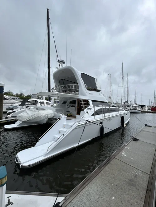 A large white yacht docked at a marina with other boats in the background under cloudy skies.