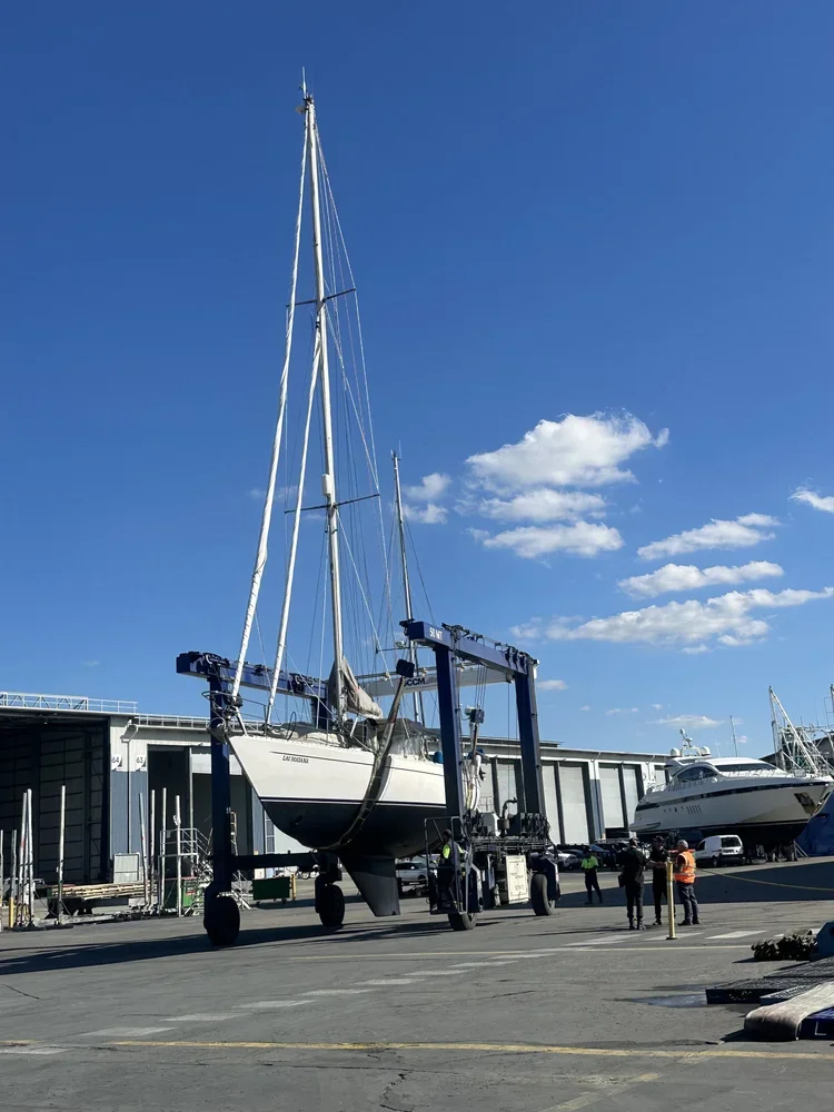 A sailboat being lifted out of the water by a boat hoist at a marina on a clear day with blue sky and scattered clouds.