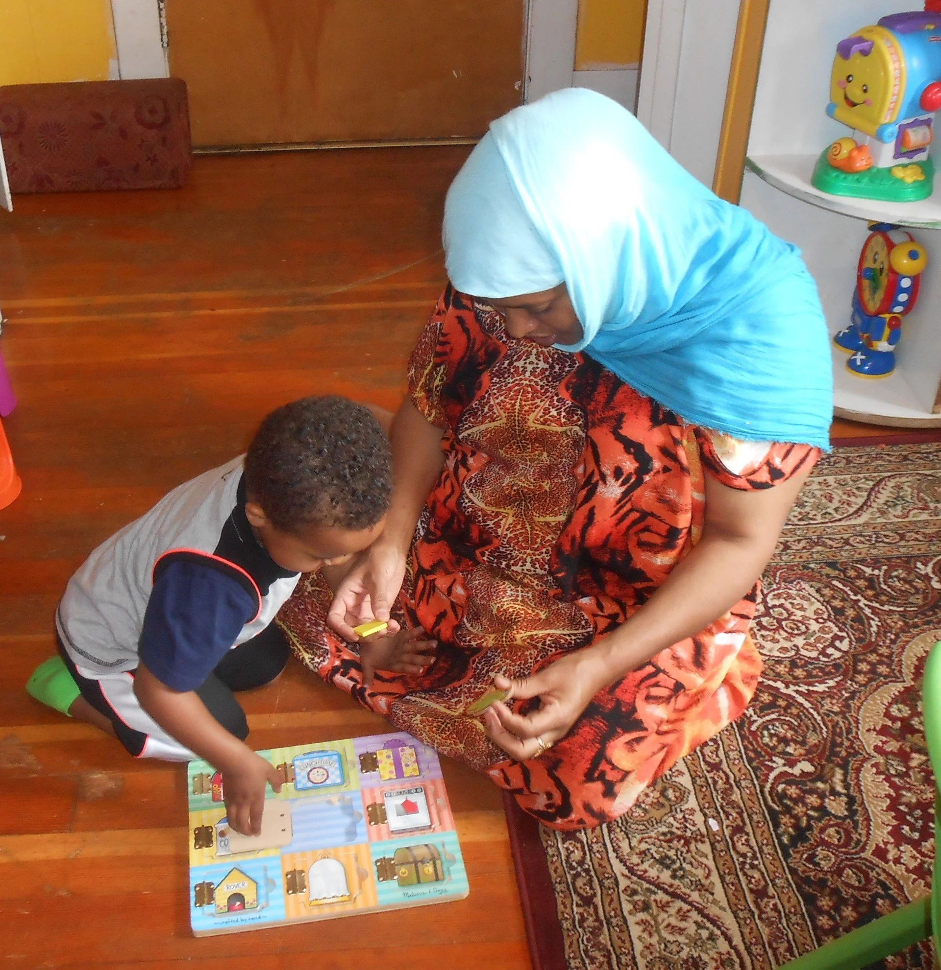 A woman wearing a light blue hijab and a red animal print skirt sits on the floor with two children, playing a colorful educational board game. The setting is indoors with a patterned rug, wooden floor, and a corner of a white cabinet with toys on it.
