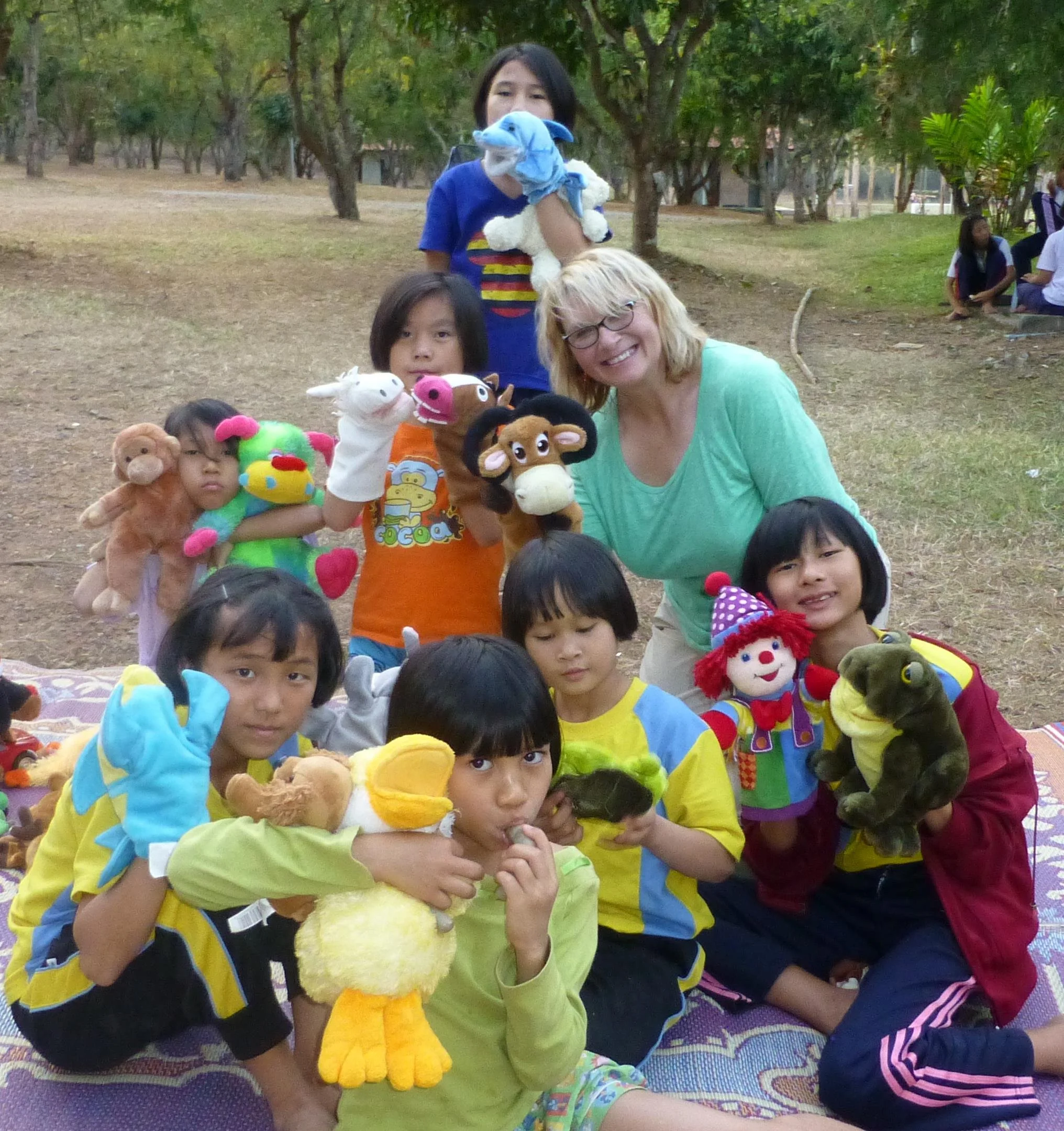 A group of children and two adults, one with glasses and blonde hair, sitting and standing outdoors on a blanket, holding puppets including animals, in a park with trees and other children in the background.