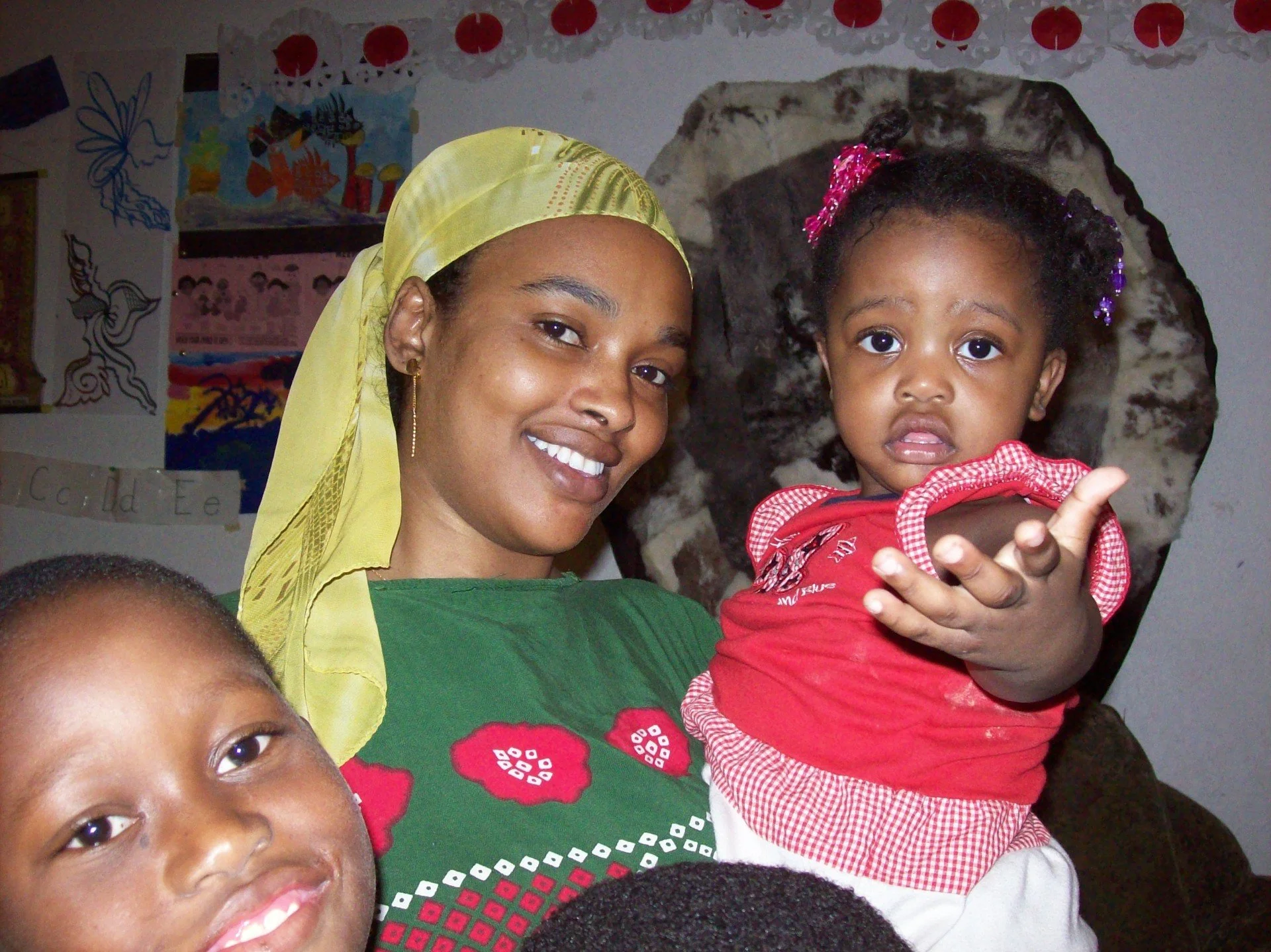 A woman with a yellow head covering and a green shirt holding a young girl with curly hair in a red shirt with white patterns. The background has colorful posters and wall decorations.