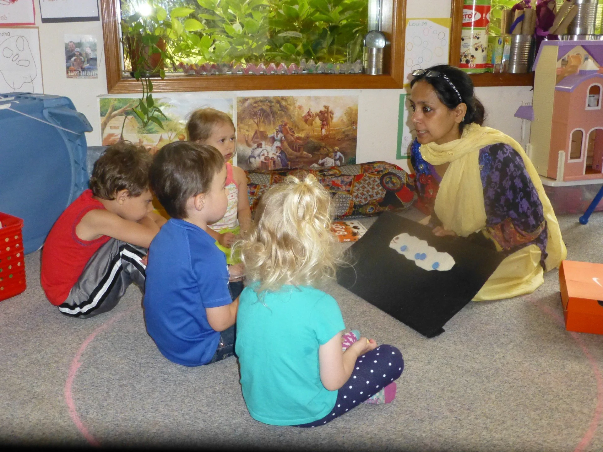 A woman teaching a group of young children in a classroom, seated on the floor, with artwork and toys in the background.