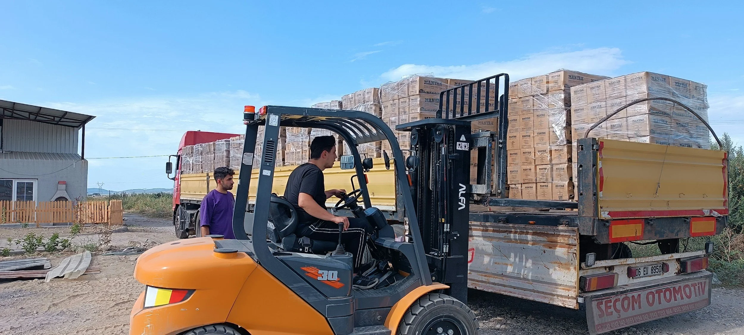 Dos personas operan un montacargas naranja que transporta cajas de cartón apiladas en un camión de carga. Otro hombre observa desde el lado del camión, en un entorno rural con estructuras de metal y un cielo despejado.