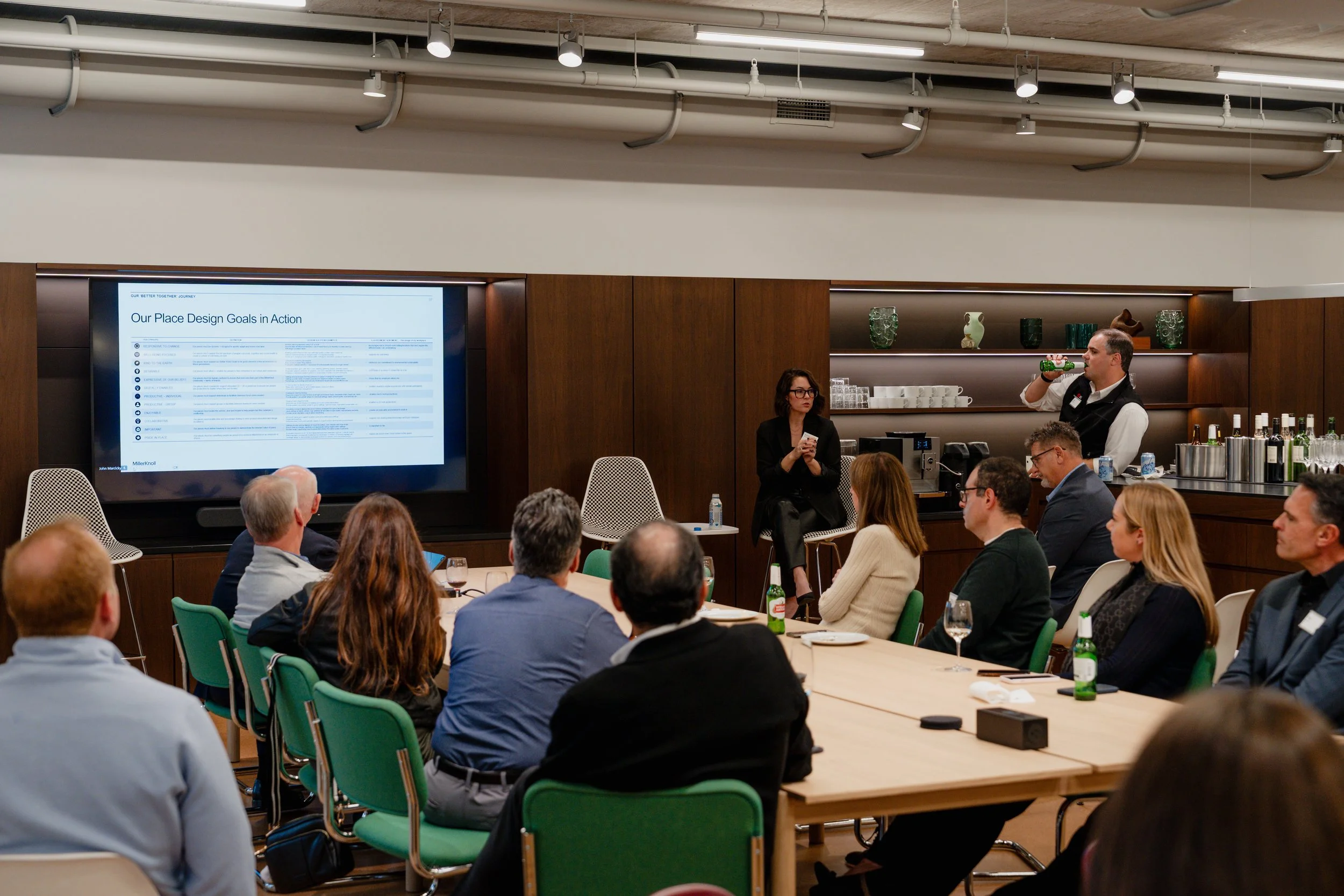 A group of people attending a conference or meeting in a modern conference room with wooden wall panels and a large screen displaying a presentation titled 'Our Place Design Goals in Action'. A man is drinking from a green bottle while a woman is standing, holding a phone. Several attendees are seated around tables, listening and paying attention.