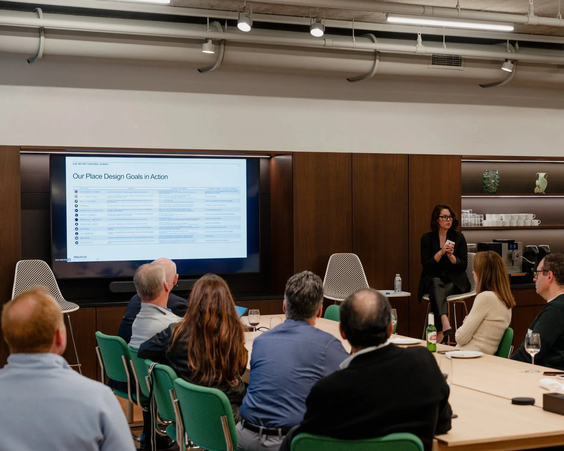 A woman in black jacket and glasses standing at the front of a room giving a presentation to an audience. A large screen behind her displays a slide titled 'Our Place Design Goals in Action.' The audience is seated around tables with some attendees appearing to listen attentively, and some with drinks on the tables.