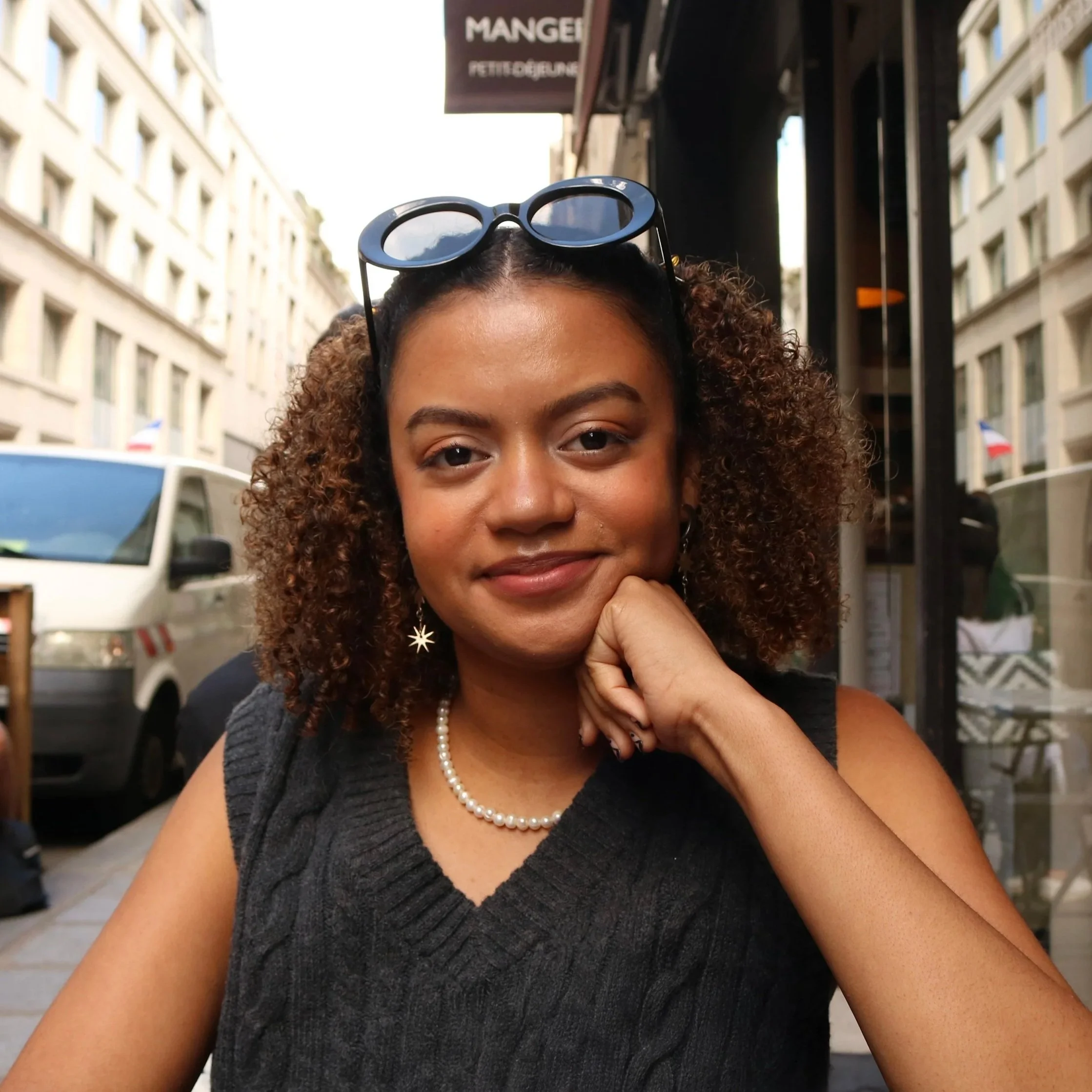 headshot of Camille Gomera-Tavarez, curly hair, wearing sunglasses outside of a Parisian cafe